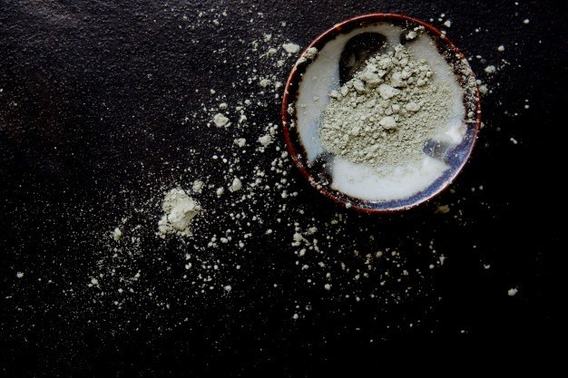 Greenish powdered cosmetic clay in a ceramic bowl on a dark surface, with powder scattered around it.