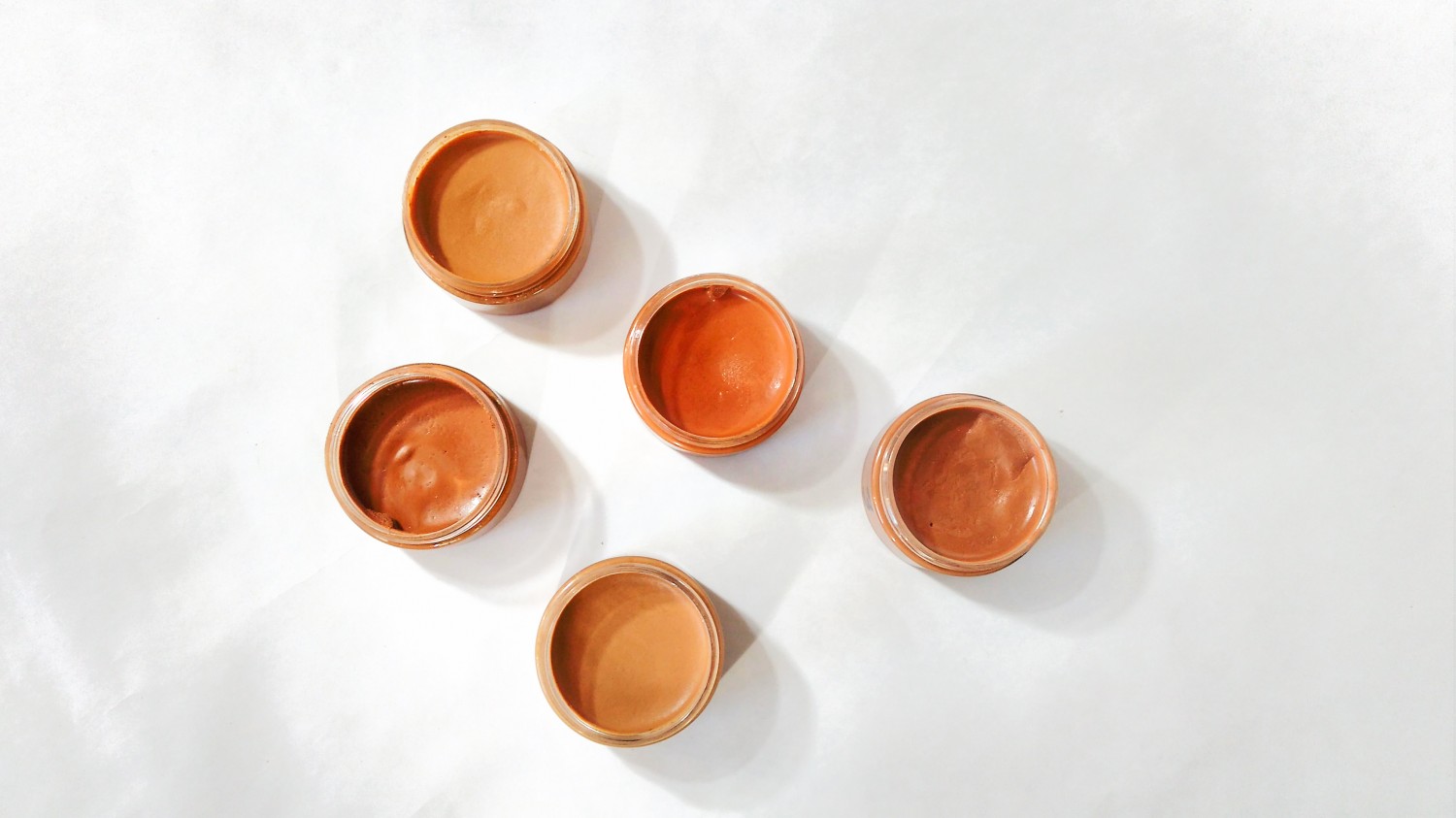 Fond de teint minéral crémeux en cinq teintes différentes, dans des pots en plastique avec couvercle en aluminium, posés sur un fond blanc.