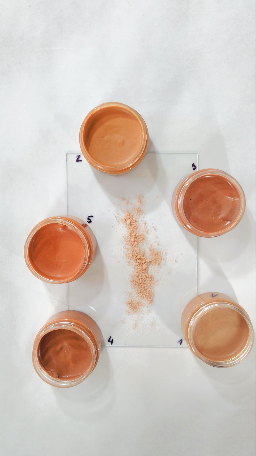Fond de teint minéral crémeux dans différentes teintes de peau, conditionné dans un pot en plastique, posé sur une plaque acrylique et un fond blanc.