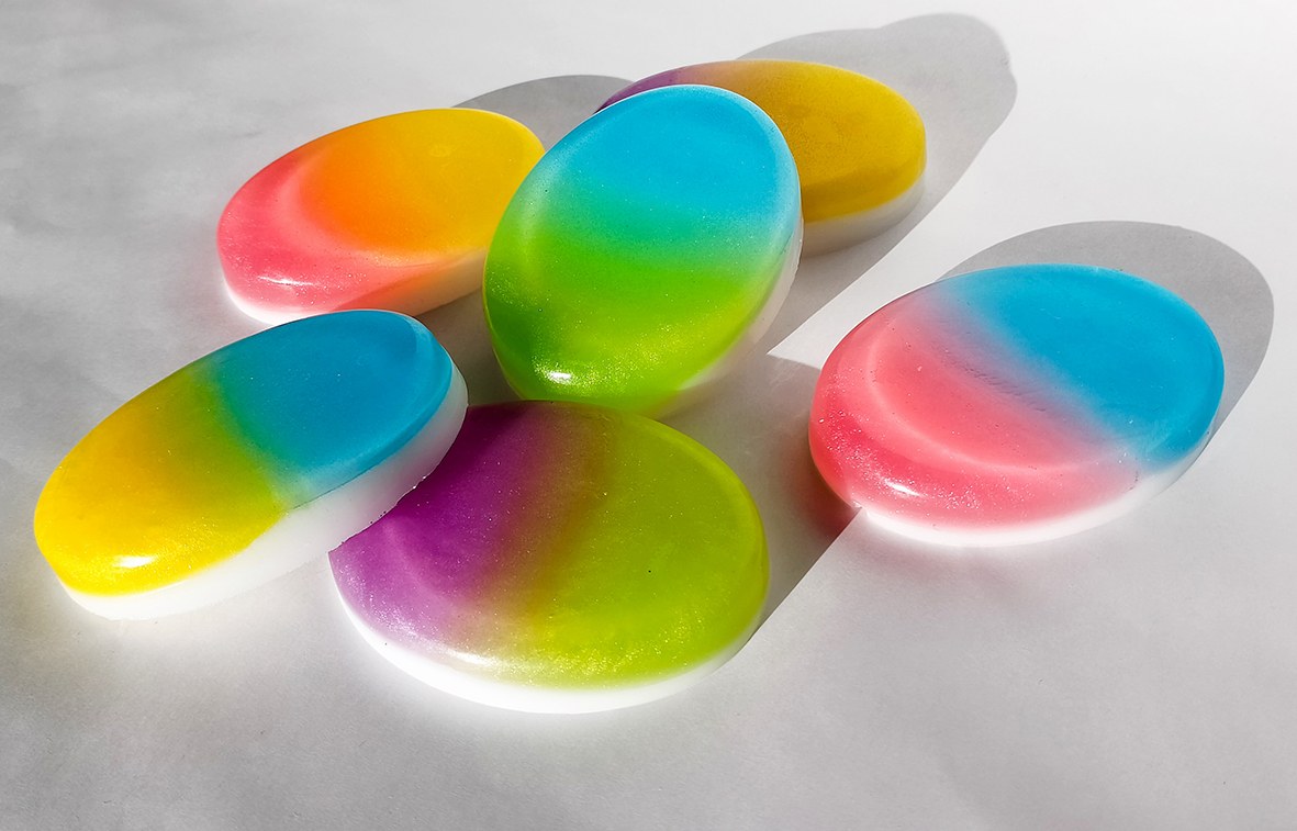 Several oval bars of ombré soap with a colour gradient and a white layer at the bottom, laid out on a white background.