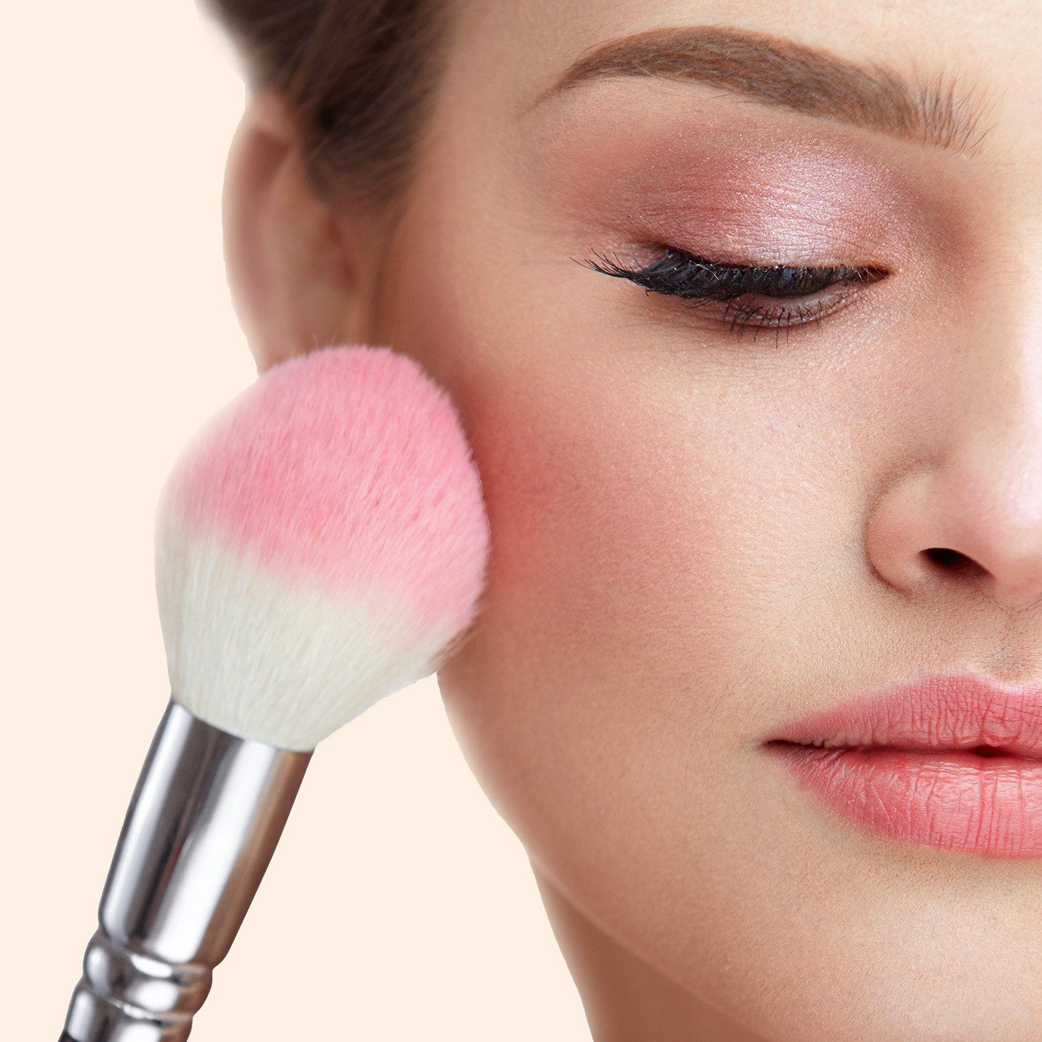 Make a blush with us in whatever colour you like. A woman applying pink blush to her face with a wide white brush.