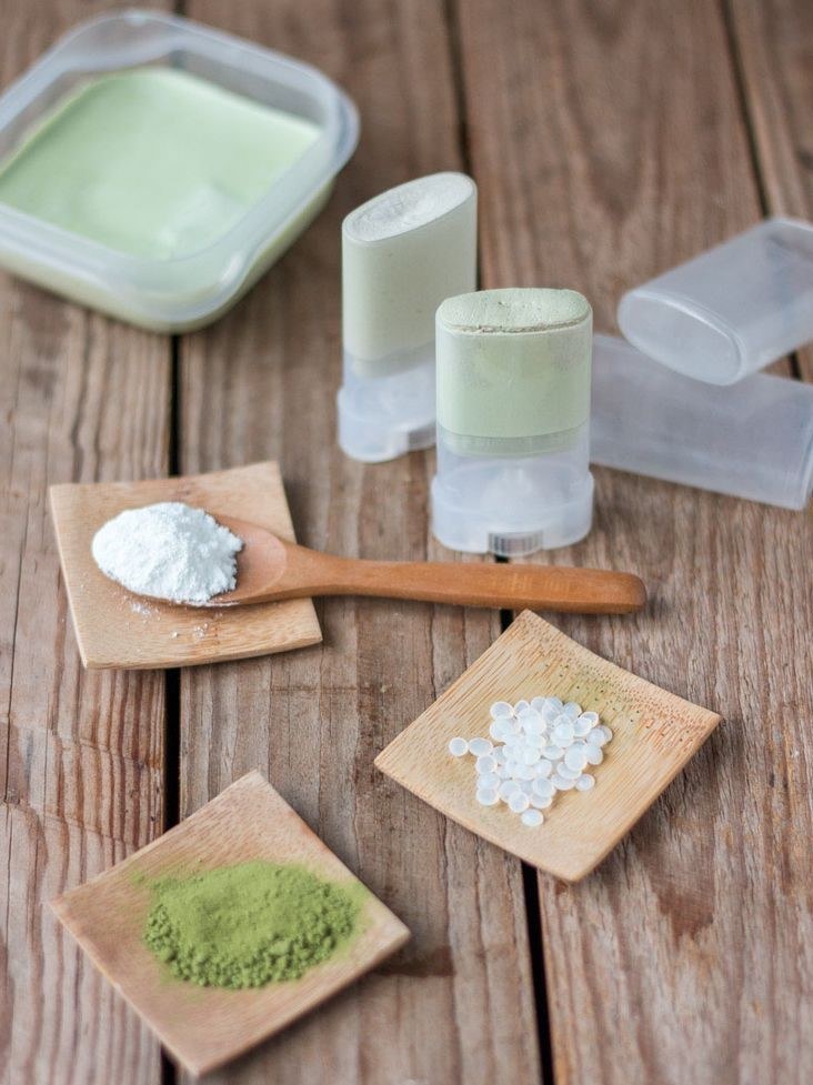 Two filled deodorant sticks and a jar of the solid mixture on a wooden table, with white and green powder and wax pastilles around them.